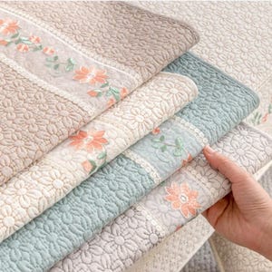 以下が含まれることがあります： パステルカラーの様々な花柄のキルトブランケットの山。各ブランケットは、詳細なデイジーパターンと、ピーチとグリーンの色合いの刺繍の花が付いた装飾的なボーダーが特徴です。ブランケットは柔らかく、魅力的です。