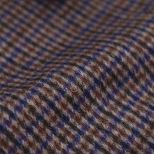 Op de afbeelding: Close-up van een textiel met een pied-de-poule patroon. De stof heeft een mix van bruine, blauwe en beige kleuren, wat een klassieke en verfijnde uitstraling creëert. De weving is zichtbaar, wat diepte en detail aan het textiel toevoegt.