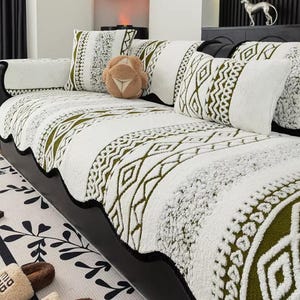 Könnte beinhalten: Eine weiß-olivgrüne Sofabezug mit Muster und passenden Kissen. Der Bezug weist geometrische Designs und Streifen auf. Ein braunes Zierkissen ist ebenfalls vorhanden. Das Sofa ist schwarz.