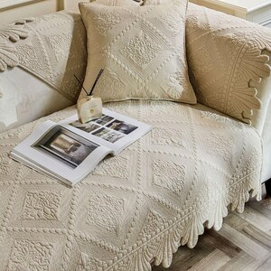 May include: Beige quilted sofa cover and pillow set with intricate floral and geometric patterns. The cover features a scalloped edge and is paired with a decorative pillow. An open magazine and a fragrance diffuser are also on the sofa.