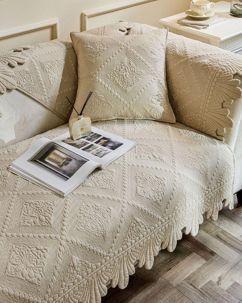 May include: Beige quilted sofa cover and pillow set with intricate floral and geometric patterns. The cover features a scalloped edge and is paired with a decorative pillow. An open magazine and a fragrance diffuser are also on the sofa.