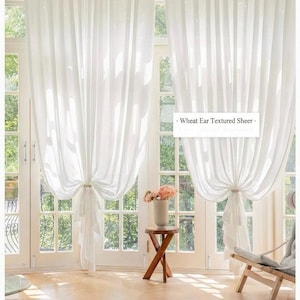 May include: Sheer white curtains with a wheat ear textured design, tied back with fabric. The curtains frame a bright window with a view of greenery. Text on the image reads: Wheat Ear Textured Sheer.