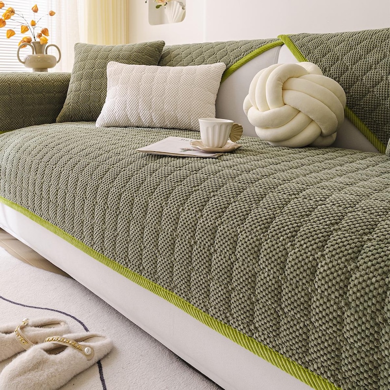 K&ouml;nnte beinhalten: Eine gesteppte Sofahusse in Salbeigr&uuml;n mit limettengr&uuml;ner Paspel. Das Sofa ist mit dekorativen Kissen in Creme und Salbeigr&uuml;n geschm&uuml;ckt. Ein cremefarbenes Knotenkissen und eine Teetasse auf einer Untertasse sind ebenfalls zu sehen.