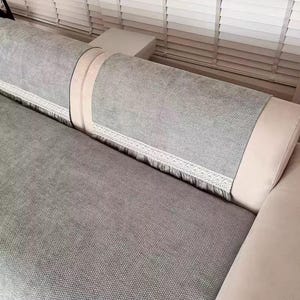 以下が含まれることがあります： テクスチャード加工された生地のライトグレーのソファ。ソファにはライトベージュのトリムと白いレースの装飾が施されています。ソファは白いブラインドの前にあります。