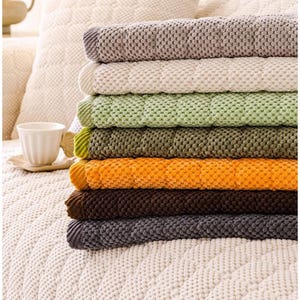 K&ouml;nnte beinhalten: Ein Stapel von sieben strukturierten Decken in verschiedenen Farben, darunter Grau, Creme, Gr&uuml;n, Olive, Orange, Braun und Dunkelgrau. Die Decken haben ein gestepptes Muster und sind ordentlich gefaltet, wodurch eine Reihe warmer und erdiger T&ouml;ne gezeigt wird.