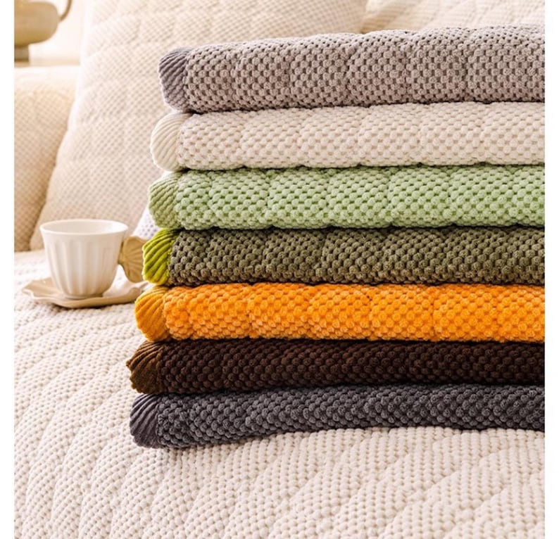 K&ouml;nnte beinhalten: Ein Stapel von sieben strukturierten Decken in verschiedenen Farben, darunter Grau, Creme, Gr&uuml;n, Olive, Orange, Braun und Dunkelgrau. Die Decken haben ein gestepptes Muster und sind ordentlich gefaltet, wodurch eine Reihe warmer und erdiger T&ouml;ne gezeigt wird.