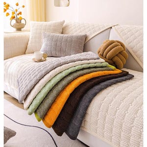 K&ouml;nnte beinhalten: Eine Auswahl an gestrickten Decken in verschiedenen Farben, darunter Grau, Gr&uuml;n, Orange und Braun, ist auf einem cremefarbenen Sofa drapiert. Die Decken haben eine strukturierte Optik. Dekorative Kissen und ein knotenf&ouml;rmiges Kissen sind ebenfalls zu sehen.