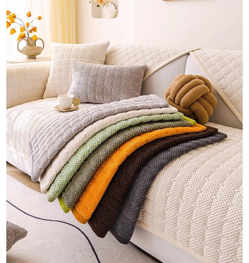 K&ouml;nnte beinhalten: Eine Auswahl an gestrickten Decken in verschiedenen Farben, darunter Grau, Gr&uuml;n, Orange und Braun, ist auf einem cremefarbenen Sofa drapiert. Die Decken haben eine strukturierte Optik. Dekorative Kissen und ein knotenf&ouml;rmiges Kissen sind ebenfalls zu sehen.