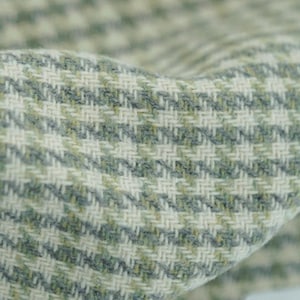 May include: Close-up of a textured fabric with a houndstooth pattern. The fabric features a mix of light green, grey, and off-white threads woven together. The weave creates a diagonal striped effect, with the fabric folded over in the image.