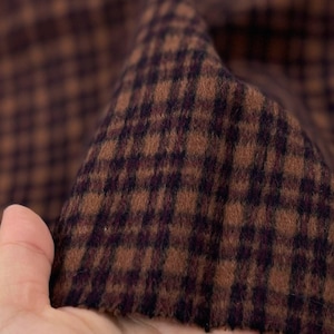 May include: Close-up of a brown and purple houndstooth fabric. The fabric is folded, revealing the texture and pattern. The material appears to be a woven wool or wool blend, suitable for apparel or home decor projects.
