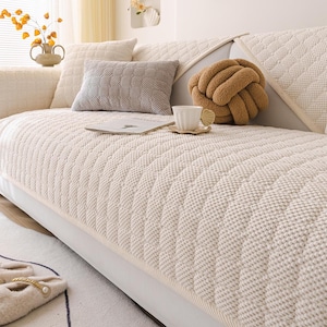 K&ouml;nnte beinhalten: Ein cremefarbenes Sofa mit einem strukturierten, gesteppten Bezug. Dekorative Kissen in Creme- und Graut&ouml;nen sind auf dem Sofa angeordnet. Ein braunes, knotenf&ouml;rmiges Kissen und eine wei&szlig;e Teetasse auf einer Untertasse erg&auml;nzen die gem&uuml;tliche &Auml;sthetik.