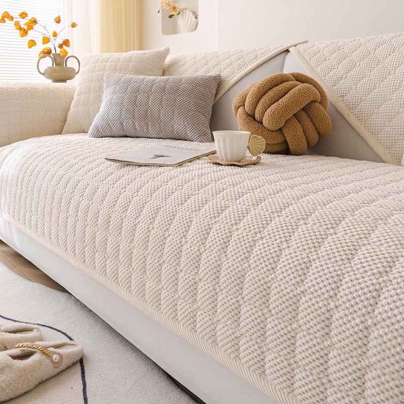 K&ouml;nnte beinhalten: Ein cremefarbenes Sofa mit einem strukturierten, gesteppten Bezug. Dekorative Kissen in Creme- und Graut&ouml;nen sind auf dem Sofa angeordnet. Ein braunes, knotenf&ouml;rmiges Kissen und eine wei&szlig;e Teetasse auf einer Untertasse erg&auml;nzen die gem&uuml;tliche &Auml;sthetik.