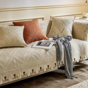 May include: A beige sofa with decorative pillows and a matching cover. The pillows are in shades of beige and rust, with intricate quilted patterns. A gray scarf and a magazine rest on the sofa.