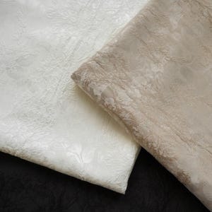 May include: Two folded fabrics with a floral pattern. One is ivory, and the other is a light beige. The fabrics have a textured, raised design. The background is black.