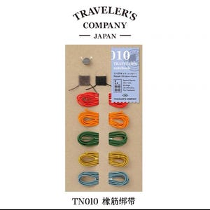 May include: A set of colourful elastic bands for a Traveler's notebook. The set includes red, orange, green, yellow, and blue bands. The bands are arranged in rows on a brown card with the text "Traveler's Company" and "TN010 橡筋绑带".