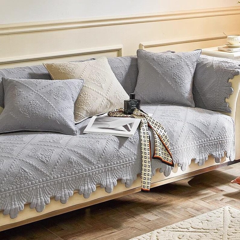 May include: A light gray quilted sofa cover with matching throw pillows. The cover has a scalloped edge and diamond pattern. Decorative pillows in light gray and cream colors are arranged on the sofa. A scarf and a perfume bottle are also on the sofa.