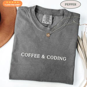 May include: A grey Comfort Colors t-shirt with the words "COFFEE & CODING" printed in white. The shirt has a crew neck and short sleeves. A silver necklace with a pendant rests on the shirt. The shirt is labelled "PEPPER".