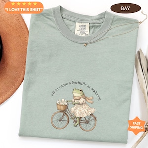 May include: Sage green Comfort Colors t-shirt featuring a frog illustration on a bicycle, wearing a dress and scarf. The text reads "off to cause a Kerfuffle at mahjong". A gold necklace and a brown hat are also visible.