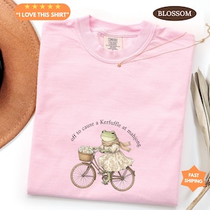 May include: A pale pink t-shirt with the text "I LOVE THIS SHIRT". The shirt features a whimsical illustration of a frog riding a bicycle with the phrase "off to cause a Kerfuffle at mahjong". The shirt has a Comfort Colors tag.