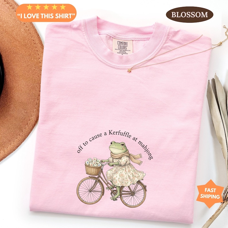 May include: A pale pink t-shirt with the text "I LOVE THIS SHIRT". The shirt features a whimsical illustration of a frog riding a bicycle with the phrase "off to cause a Kerfuffle at mahjong". The shirt has a Comfort Colors tag.