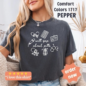 May include: Dark grey t-shirt featuring white illustrations of pills, a dropper, a lab coat, and a chemical structure. The text "will yap about pills" is below the illustrations. The shirt also has the text "Comfort Colors 1717 PEPPER".