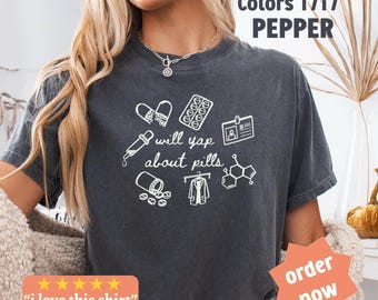 May include: Dark gray t-shirt with white graphic illustrations of pills, a dropper, a lab coat, and a chemical structure. The text "will yap about pills" is centered below the illustrations. The shirt also has the text "Comfort Colors 1717 PEPPER".