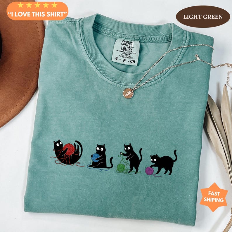 May include: Light green t-shirt with a graphic of four black cats playing with yarn in red, blue, and green. The shirt has the text "I LOVE THIS SHIRT" and "COMFORT COLORS" printed on it.
