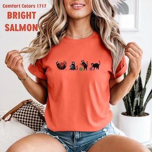 May include: Bright salmon-colored t-shirt with a graphic of four black cats playing with yarn balls. The shirt has the text "Comfort Colors 1717" and "BRIGHT SALMON" printed on the top. The shirt is a crew neck and has short sleeves.