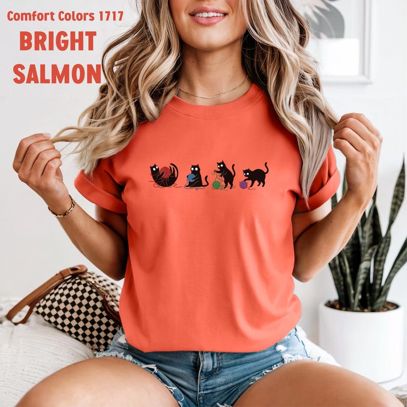 May include: Bright salmon-colored t-shirt with a graphic of four black cats playing with yarn balls. The shirt has the text "Comfort Colors 1717" and "BRIGHT SALMON" printed on the top. The shirt is a crew neck and has short sleeves.