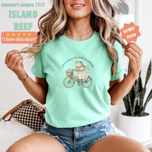 May include: A pale green Comfort Colors t-shirt with the text "ISLAND REEF" and "i love this shirt". The shirt features a frog on a bicycle with the text "off to cause a Kerfuffle at mahjong".