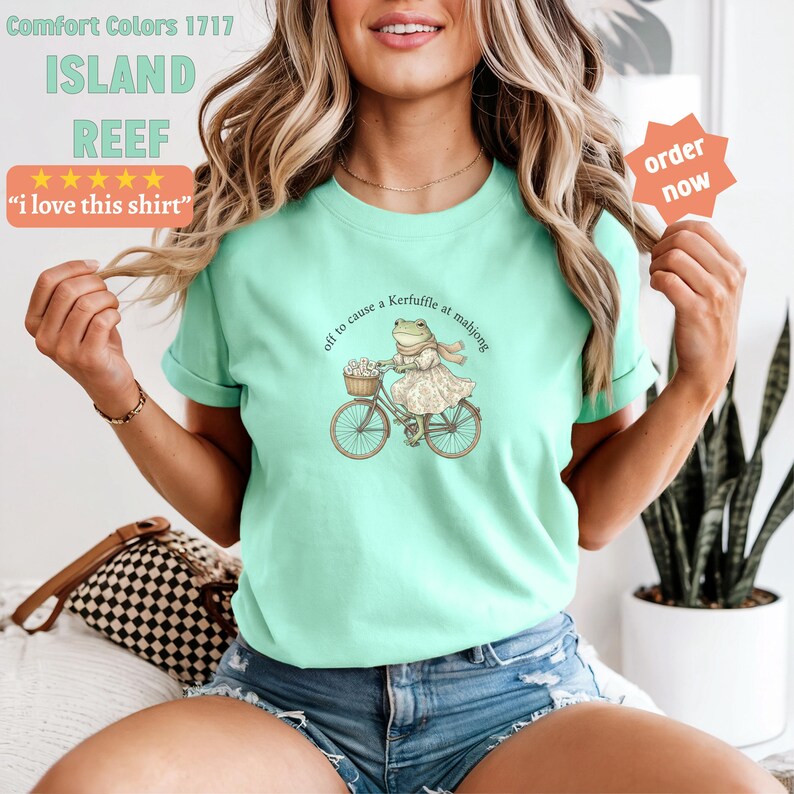 May include: A pale green Comfort Colors t-shirt with the text "ISLAND REEF" and "i love this shirt". The shirt features a frog on a bicycle with the text "off to cause a Kerfuffle at mahjong".