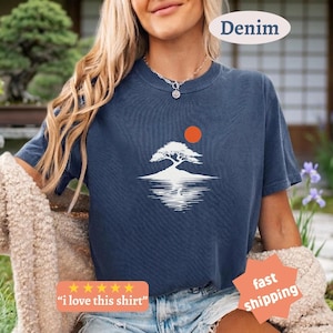May include: Denim blue t-shirt featuring a white tree graphic with a red sun and water reflection. The shirt has the text "i love this shirt" and "fast shipping" on it. The shirt is a casual style.