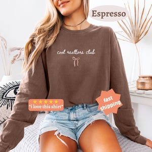 May include: A brown long-sleeve crewneck sweatshirt featuring the text "cool realtors club" in white cursive lettering above a small white line-art bow graphic. The sweatshirt is paired with light blue denim shorts with frayed edges.