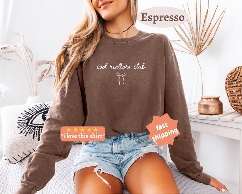 May include: A brown long-sleeve crewneck sweatshirt featuring the text "cool realtors club" in white cursive lettering above a small white line-art bow graphic. The sweatshirt is paired with light blue denim shorts with frayed edges.