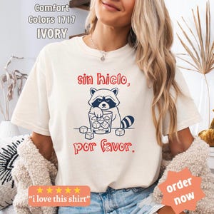 Könnte beinhalten: Elfenbeinfarbenes T-Shirt mit einer Illustration eines Waschbären, der ein Glas Eiswürfel hält, und dem spanischen Text "sin hielo, por favor." Das Shirt hat auch den Text "i love this shirt" und "order now".