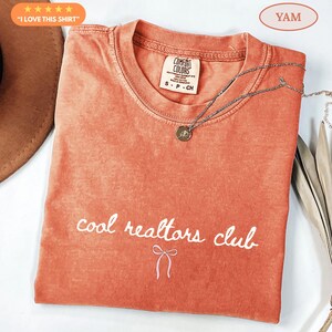 May include: A terracotta-colored short-sleeve t-shirt with a crew neckline. The shirt features the white text "cool realtors club" printed in a cursive font above a small white line-art bow graphic. A silver-toned necklace rests on the fabric.