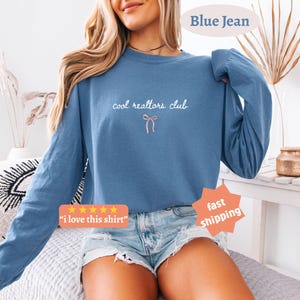 May include: A blue long-sleeve crewneck sweatshirt featuring the white text "cool realtors club" centered on the chest above a small, simple pink line-art bow graphic. The sweatshirt has a relaxed fit with ribbed cuffs and a ribbed neckline.