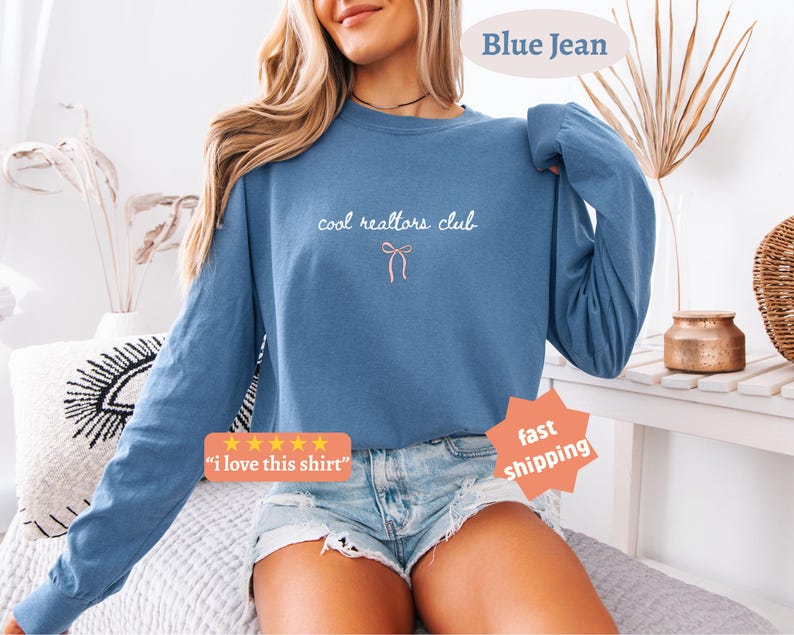 May include: A blue long-sleeve crewneck sweatshirt featuring the white text "cool realtors club" centered on the chest above a small, simple pink line-art bow graphic. The sweatshirt has a relaxed fit with ribbed cuffs and a ribbed neckline.