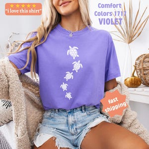 May include: A violet-colored t-shirt with a white turtle design. The shirt has the text "Comfort Colors 1717 VIOLET" and "I love this shirt" on it. The model is wearing denim shorts and a beige cardigan.