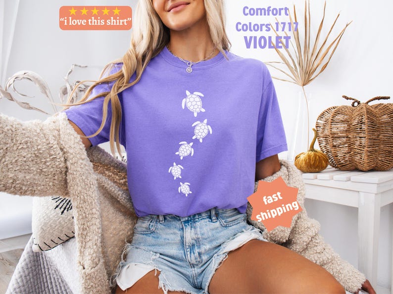 May include: A violet-colored t-shirt with a white turtle design. The shirt has the text "Comfort Colors 1717 VIOLET" and "I love this shirt" on it. The model is wearing denim shorts and a beige cardigan.
