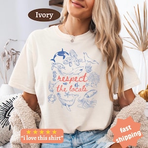 Könnte beinhalten: Elfenbeinfarbenes T-Shirt mit einem Design zum Thema Meereslebewesen. Das Shirt zeigt blaue Illustrationen verschiedener Meeresbewohner, darunter ein Oktopus, ein Wal und ein Hai, die den Text "respect the locals" in Rot und Blau umgeben. Das Shirt hat auch den Text "I love this shirt".