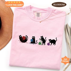May include: Pink Comfort Colors t-shirt with a graphic of four black cats playing with yarn. The text "I LOVE THIS SHIRT" is in a yellow banner at the top left. The shirt has a crew neck and short sleeves.