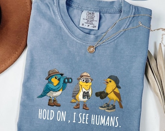 May include: A blue Comfort Colors t-shirt with a graphic of three cartoon birds wearing hats and holding binoculars. The text "HOLD ON, I SEE HUMANS" is printed below the birds. The shirt also has the text "BLUE JEAN" printed on it.