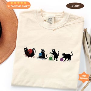 May include: Ivory-colored t-shirt featuring four black cat silhouettes playing with colorful yarn balls. The shirt has a crew neck and a Comfort Colors tag. Text on the shirt says "I LOVE THIS SHIRT".
