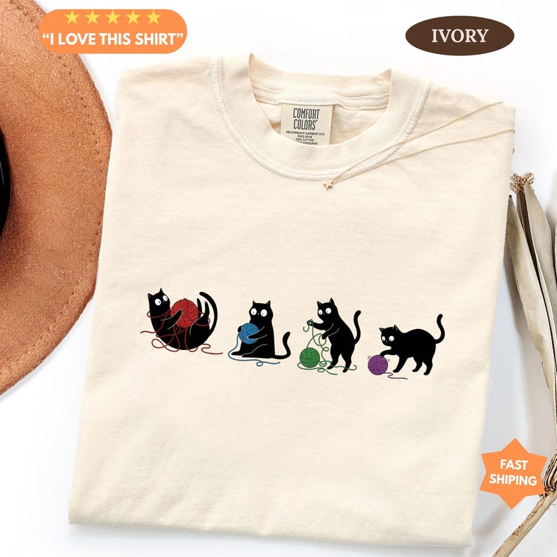 May include: Ivory-colored t-shirt featuring four black cat silhouettes playing with colorful yarn balls. The shirt has a crew neck and a Comfort Colors tag. Text on the shirt says "I LOVE THIS SHIRT".