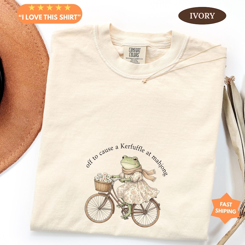 May include: Cream-coloured t-shirt featuring a frog on a bicycle, with mahjong tiles in the basket. The text says "off to cause a Kerfuffle at mahjong." The shirt also has the text "I LOVE THIS SHIRT".