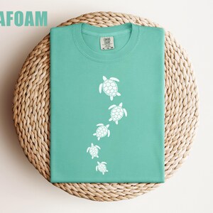 May include: Seafoam green t-shirt with a white turtle design. The shirt is folded on a woven, circular surface. The word "SEAFOAM" is printed in green at the top left of the image. The shirt has a crew neck and short sleeves.