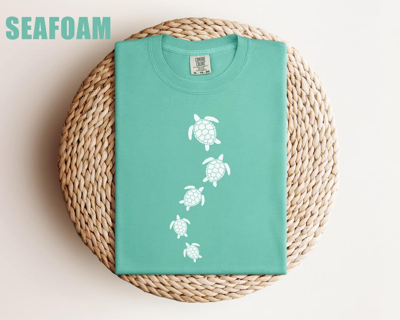 May include: Seafoam green t-shirt with a white turtle design. The shirt is folded on a woven, circular surface. The word "SEAFOAM" is printed in green at the top left of the image. The shirt has a crew neck and short sleeves.