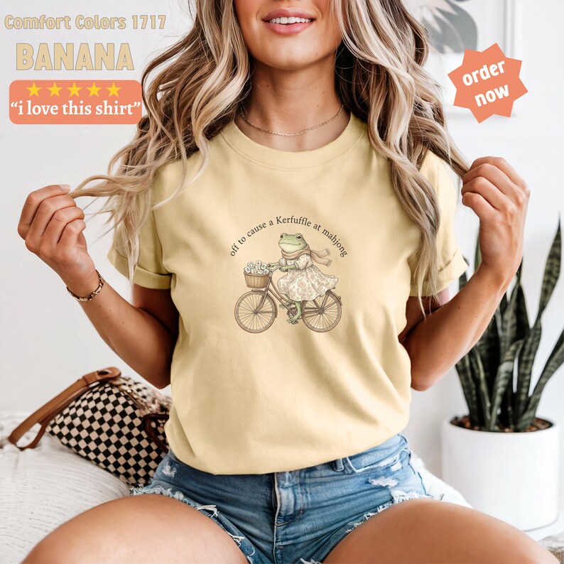 May include: Pale yellow t-shirt featuring a frog on a bicycle, with the phrase "off to cause a Kerfuffle at mahjong". The shirt also has the text "i love this shirt".