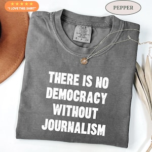 Op de afbeelding: Een antracietgrijs t-shirt met korte mouwen en de witte, verweerde tekst "THERE IS NO DEMOCRACY WITHOUT JOURNALISM" op de voorzijde. Het shirt heeft een ronde hals en een Comfort Colors-merklabel aan de binnenkant van de kraag.